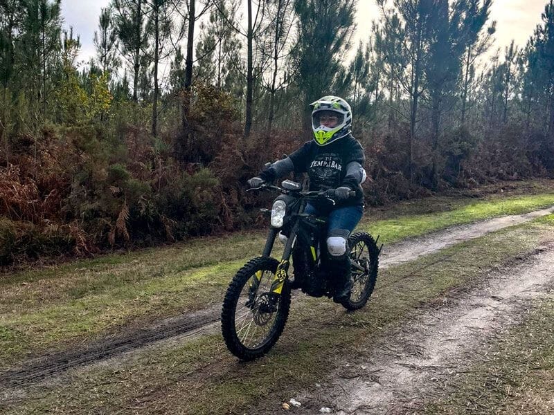 Personne en tenue de protection roulant une moto électrique tout-terrain sur un chemin forestier en pleine nature.