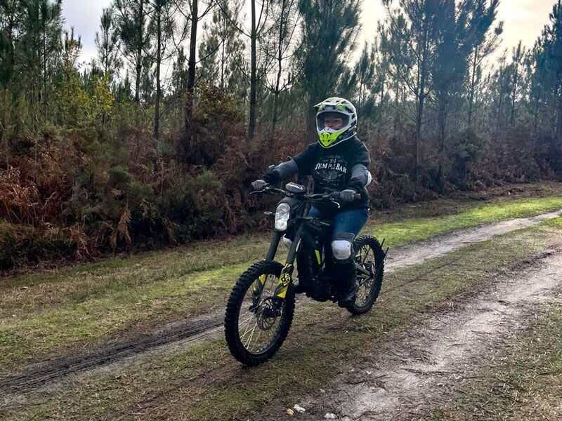 Personne en tenue de protection roulant une moto électrique tout-terrain sur un chemin forestier en pleine nature.