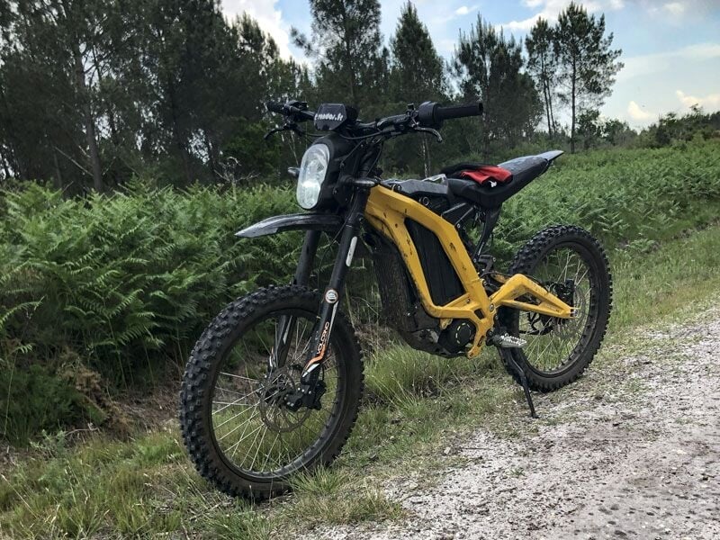 Moto électrique tout-terrain noire et jaune stationnée sur un chemin de gravier entouré de fougères et d'arbres.