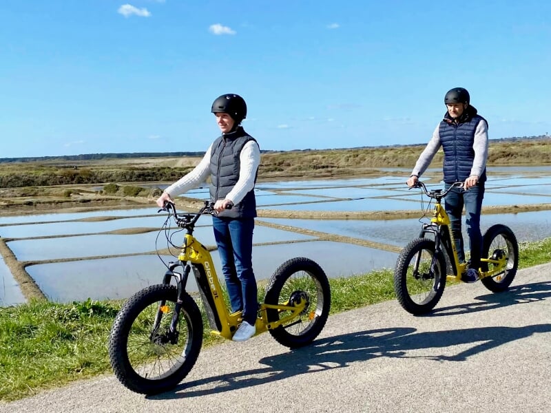 Deux personnes portant un casque et un gilet sans manches roulent sur des trottinettes électriques jaunes sur une route près des marais salants.