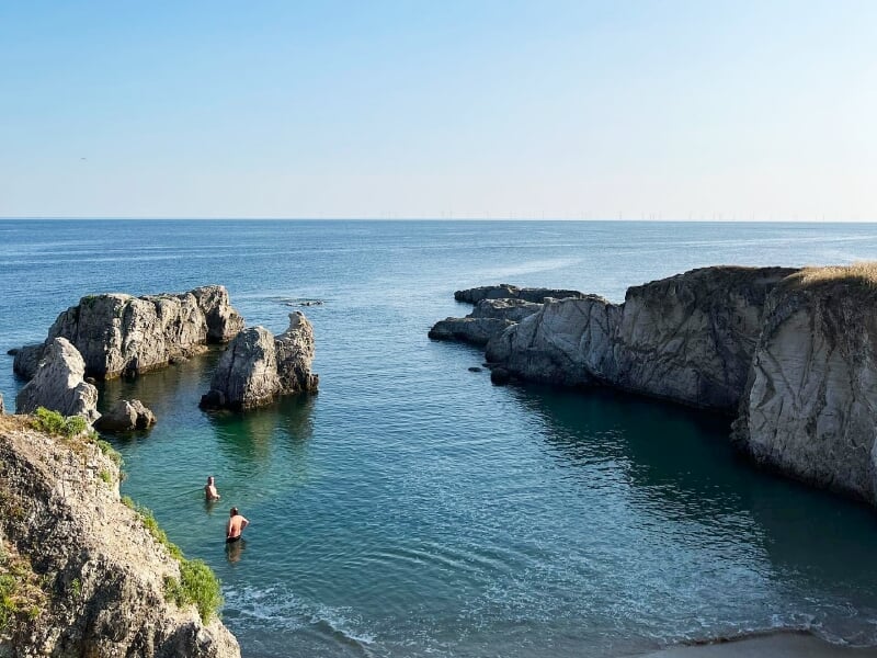 Vue sur une crique rocheuse avec deux personnes dans l'eau, au bord de la mer calme sous un ciel clair.