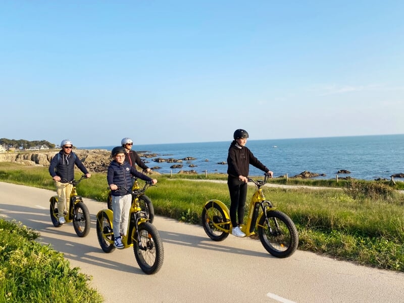 Quatre personnes portant des casques roulent en trottinettes électriques jaunes sur une piste au bord de la mer au Pouliguen.