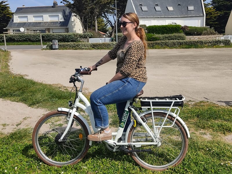 Femme souriante assise sur un vélo électrique blanc dans un cadre verdoyant avec maisons en arrière-plan.