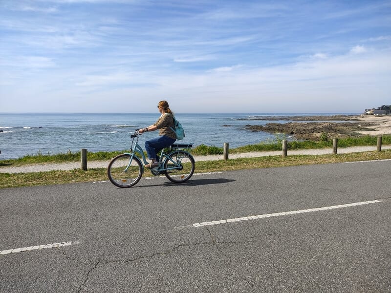 Femme en jean et pull marron roulant à vélo électrique bleu le long d'une route près de la mer et des rochers.