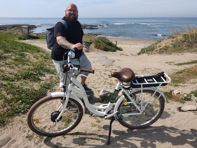 Homme barbu avec sac à dos debout à côté d'un vélo électrique blanc sur une plage sableuse avec l'océan en arrière-plan.
