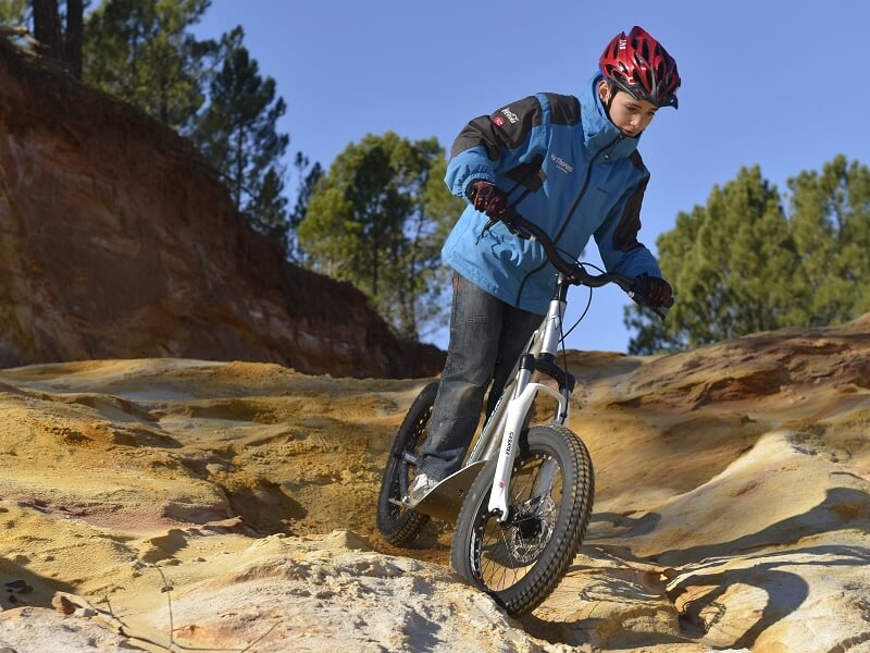 Jeune garçon en casque rouge et veste bleue descend un terrain rocheux accidenté sur une trottinette électrique tout-terrain.