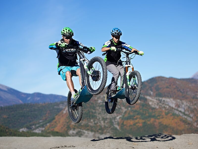 Deux personnes portant casque et gants font un saut en trottinette électrique tout-terrain dans un paysage montagneux clair.