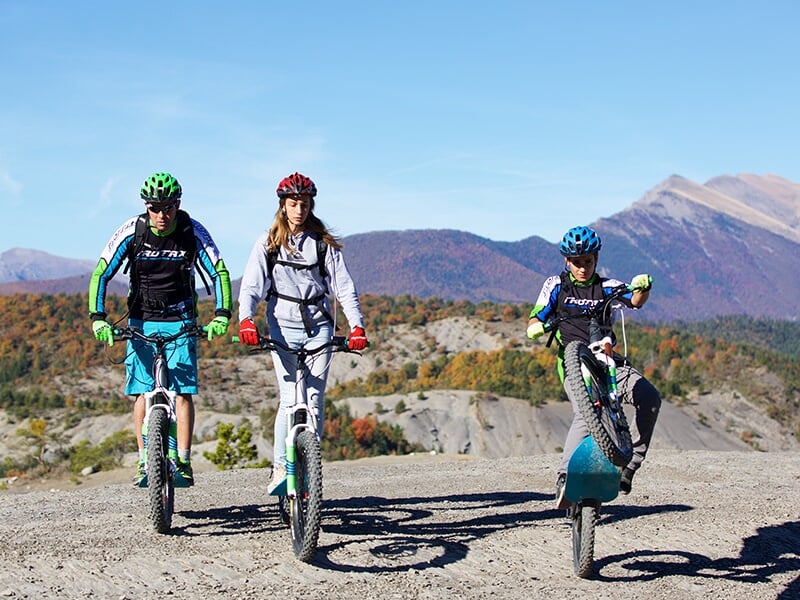 Trois personnes en tenue de sport et casque font du vélo électrique tout terrain sur un chemin de montagne ensoleillé.