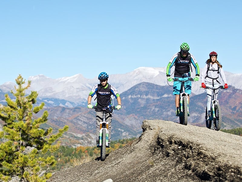 Trois personnes en tenue de sport font du vélo électrique sur un sentier montagneux avec un paysage de montagnes en arrière-plan.