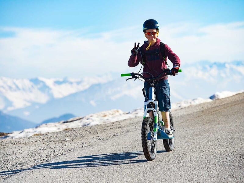 Enfant portant un casque et des lunettes de soleil, faisant un signe de paix en roulant sur une trottinette électrique en montagne.
