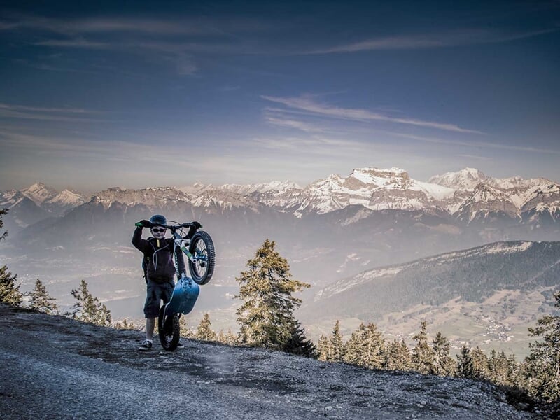 Personne en tenue de sport effectuant une roue avant avec une trottinette électrique sur une route de montagne enneigée.
