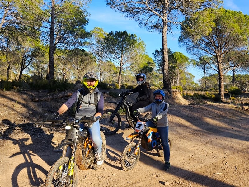 Trois motards en casque posant avec leurs vélos tout-terrain dans une zone forestière pittoresque, prêts pour une balade aventureuse.