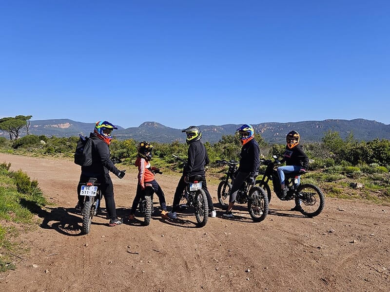 Un groupe de cinq motards sur des vélos tout-terrain électriques, se faisant face sur un chemin de terre avec une vue panoramique sur les montagnes et un ciel bleu en arrière-plan.