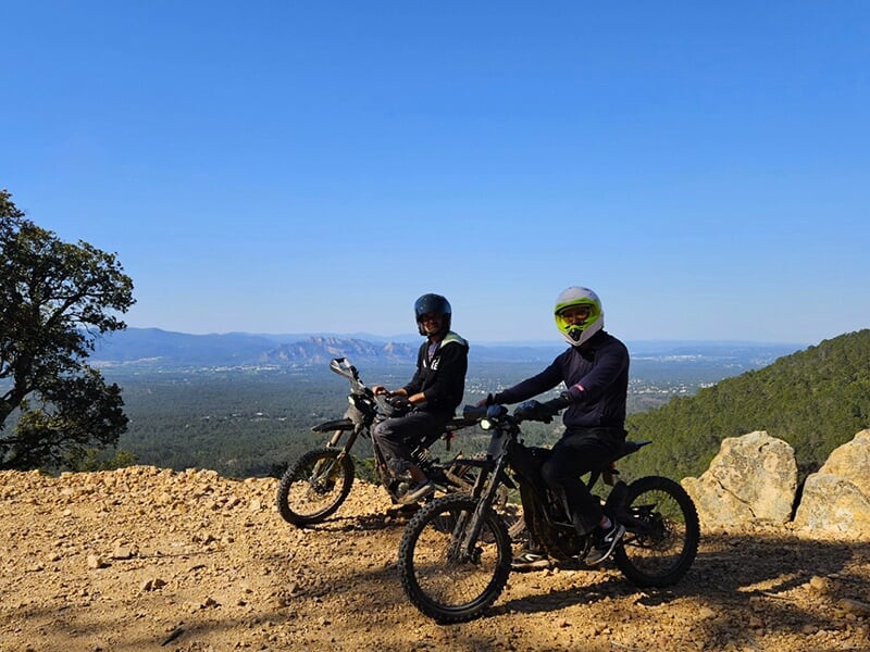 Deux motards en casques, assis sur leurs vélos tout-terrain à un point de vue rocheux et élevé, avec des vues étendues sur le paysage et les montagnes.