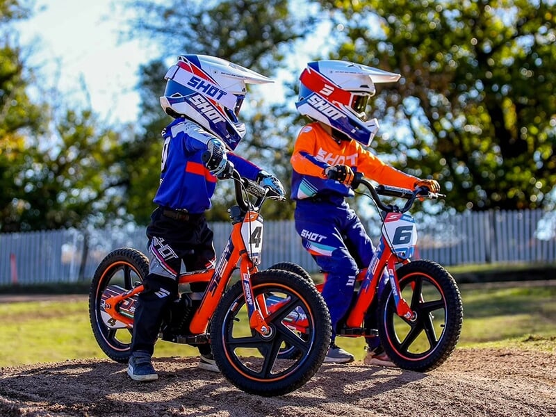 Deux jeunes enfants en tenue de protection et casques, debout côte à côte avec leurs petits vélos orange, prêts pour une balade sur une piste en terre.