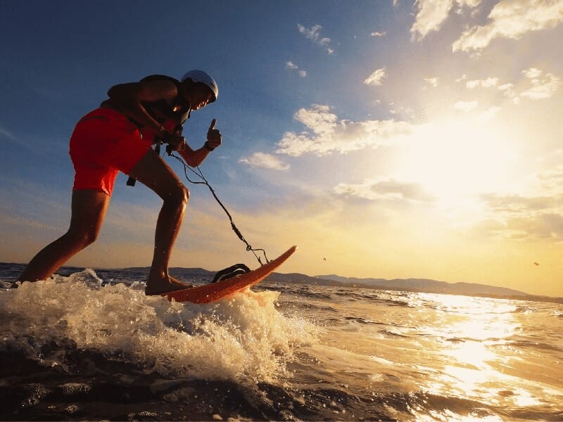 Personne faisant du surf électrique au coucher du soleil, avec casque et gilet de sécurité, sur une mer légèrement agitée.