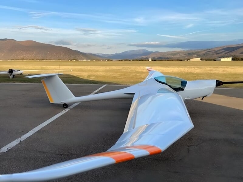 Planeur blanc avec marquages orange garé sur la piste de l'aérodrome de Château-Arnoux, montagnes en arrière-plan.