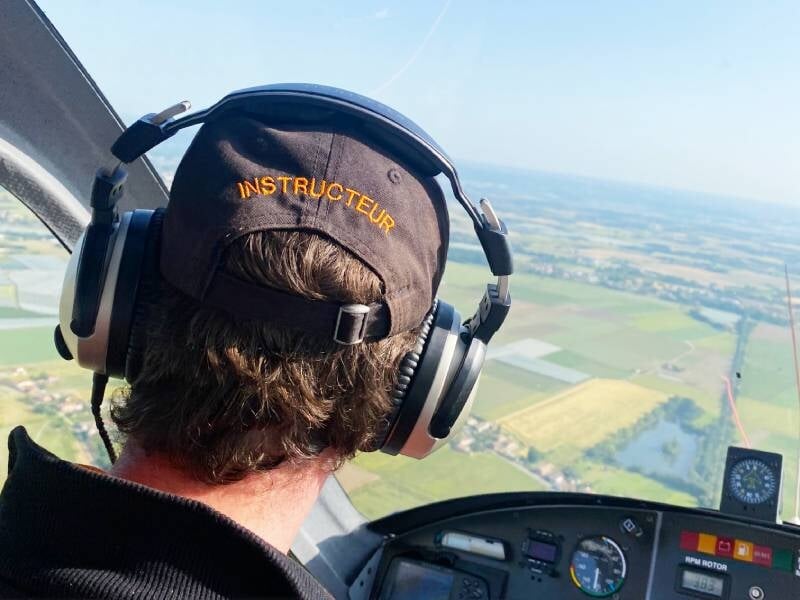 Vue depuis un cockpit d'avion avec un instructeur portant un casque audio et une casquette noire brodée en jaune.
