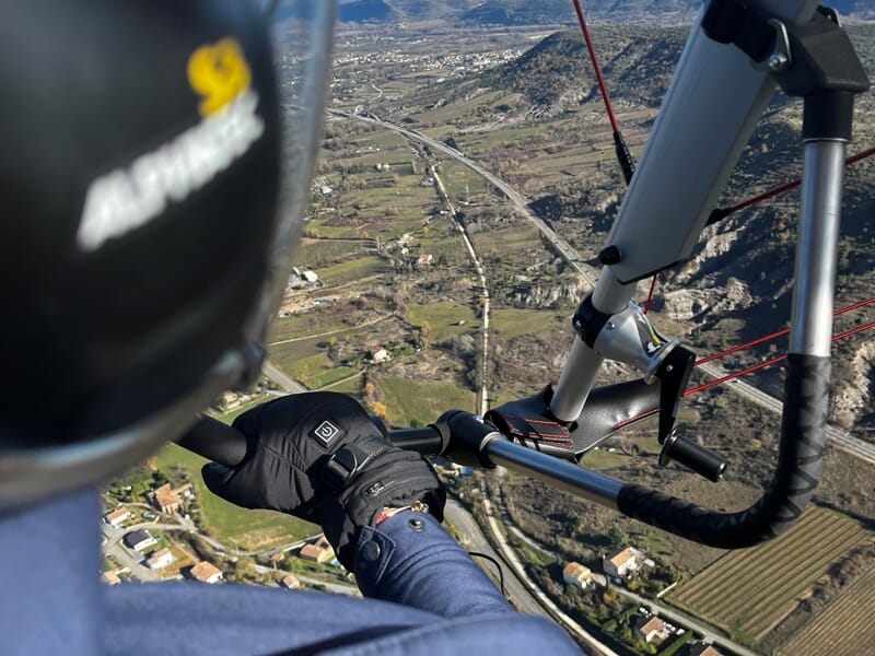 Pilote en vol dans un ULM, tenant le manche, survolant une vallée avec des champs et des collines.