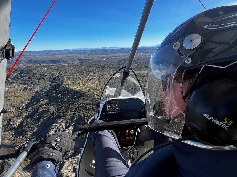Pilote équipé d'un casque dans un ULM, volant au-dessus d'un terrain montagneux sous un ciel dégagé.
