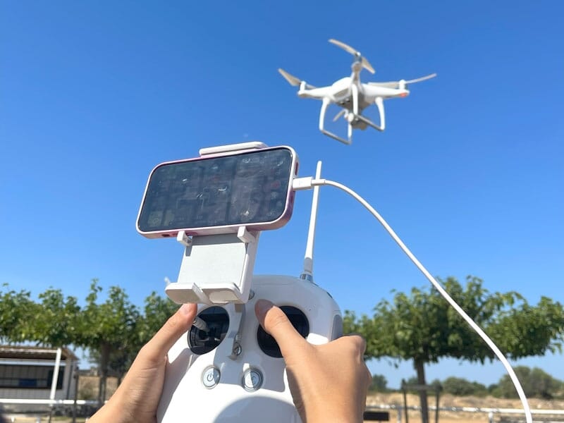 Télécommande de drone en gros plan, tenant un smartphone connecté, avec un drone blanc en vol en arrière-plan, ciel bleu et arbres