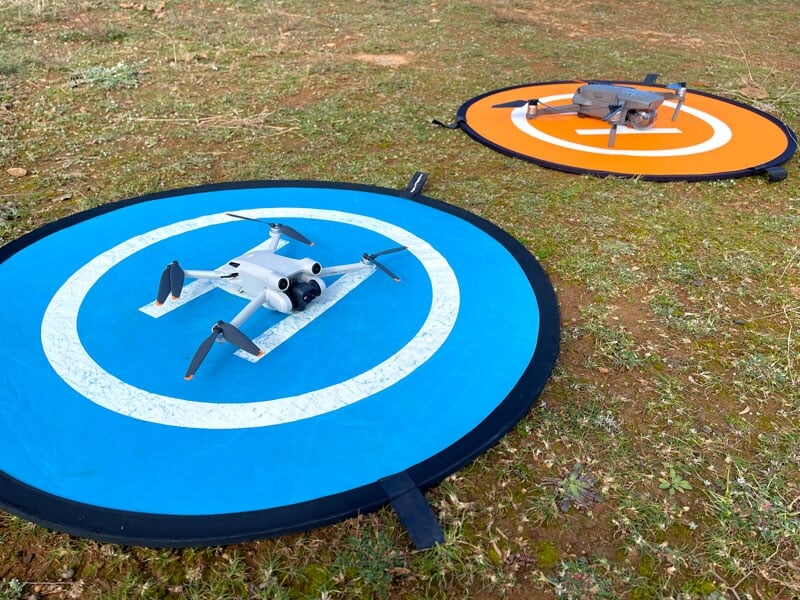 Deux drones sur des pistes d'atterrissage circulaires, l'une bleue avec un drone blanc et l'autre orange avec un drone gris, posés sur de l'herbe