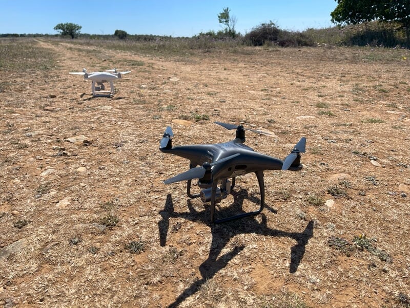 Deux drones au sol, l'un noir au premier plan et l'autre blanc en arrière-plan, sur un terrain sec et rocailleux