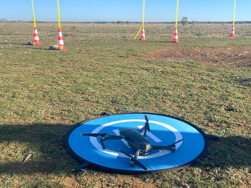 Drone noir sur une piste d'atterrissage circulaire bleue dans un champ, entouré de cônes de signalisation et de balises jaunes