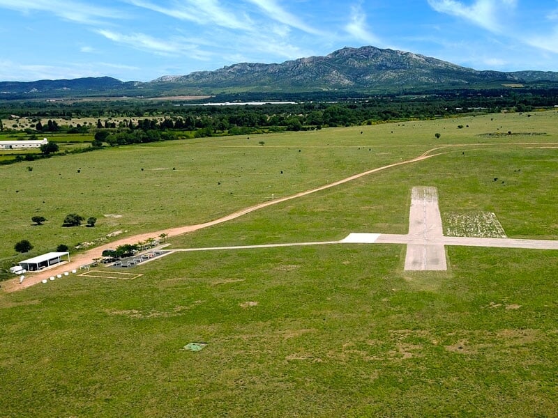 Vue aérienne d'un terrain de vol pour drones avec des pistes en béton et une grande étendue de verdure, montagnes à l'horizon sous un ciel bleu partiellement nuageux