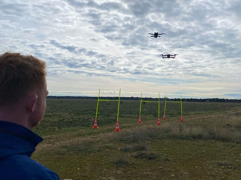 Homme regardant deux drones en vol, avec des cônes de signalisation et des balises jaunes en arrière-plan, sous un ciel couvert