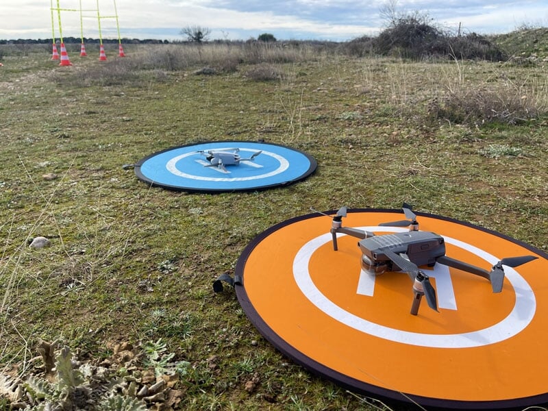 Deux drones sur des pistes d'atterrissage circulaires, l'une bleue avec un drone blanc et l'autre orange avec un drone gris, sur un terrain herbeux, cônes de signalisation en arrière-plan