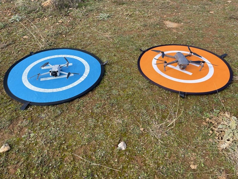 Deux drones sur des pistes d'atterrissage circulaires, l'une bleue avec un drone blanc et l'autre orange avec un drone gris, sur un terrain herbeux