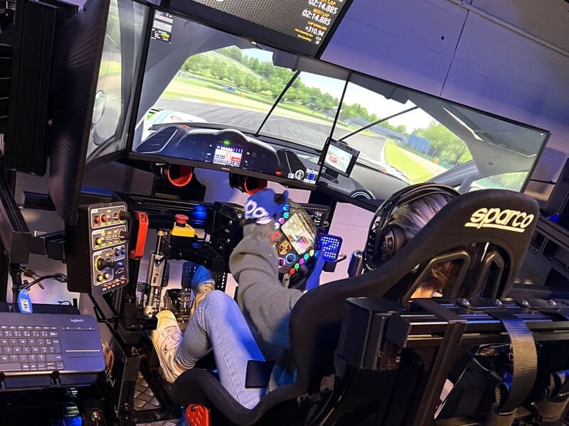 Jeune personne en pleine session de simulation de course, tenant fermement un volant à multiples boutons colorés. Les écrans montrent une vue de piste immersive. Le siège Sparco et les accessoires aut