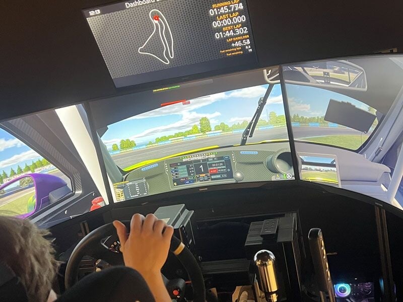 Personne utilisant un simulateur de conduite avec volant et plusieurs écrans affichant une course automobile.