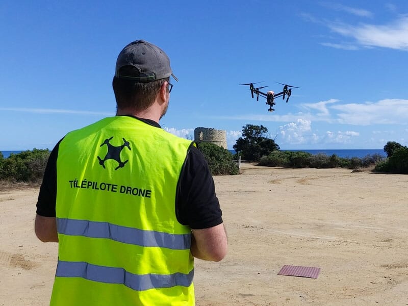 Télépilote de drone portant un gilet jaune fluorescent, contrôlant un drone en vol en extérieur près de la mer, ciel bleu dégagé