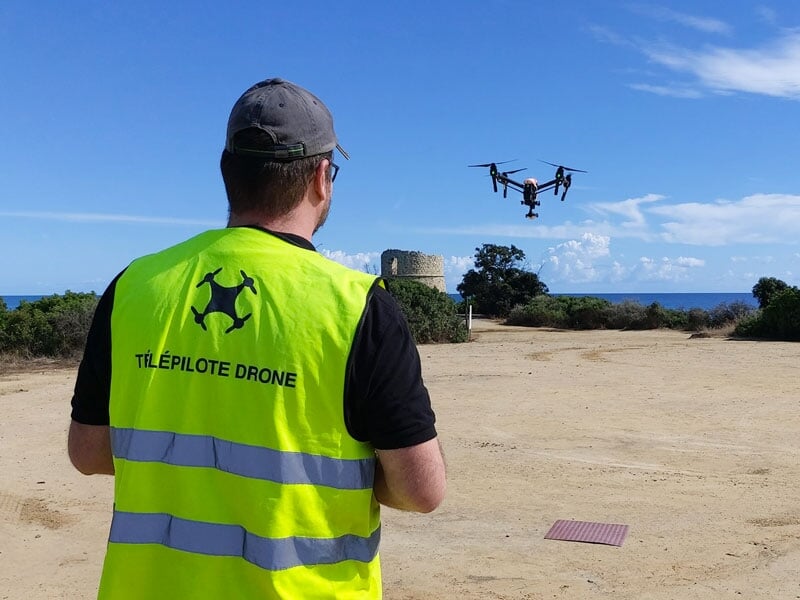 Télépilote de drone portant un gilet jaune fluorescent, contrôlant un drone en vol en extérieur près de la mer, ciel bleu dégagé