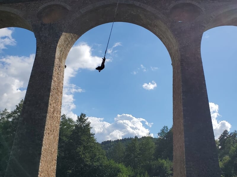 Personne suspendue en plein saut à l'élastique sous un grand viaduc en pierre avec ciel bleu et nuages.