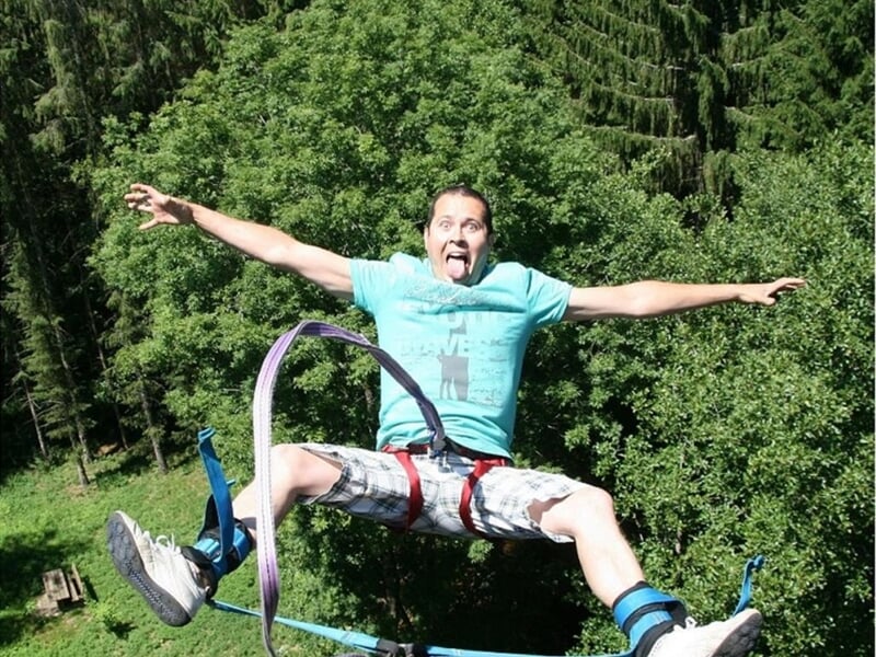 Homme en t-shirt bleu clair sautant à l'élastique depuis un viaduc en pierre entouré de forêt dense.