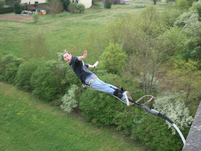 Homme en jean et t-shirt noir en plein saut à l'élastique au-dessus d'une zone herbeuse et boisée.