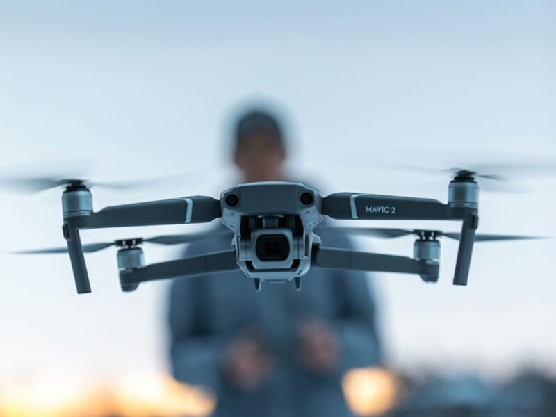 Un drone Mavic 2 en vol avec un pilote flou en arrière-plan, capturant une scène en extérieur au crépuscule.
