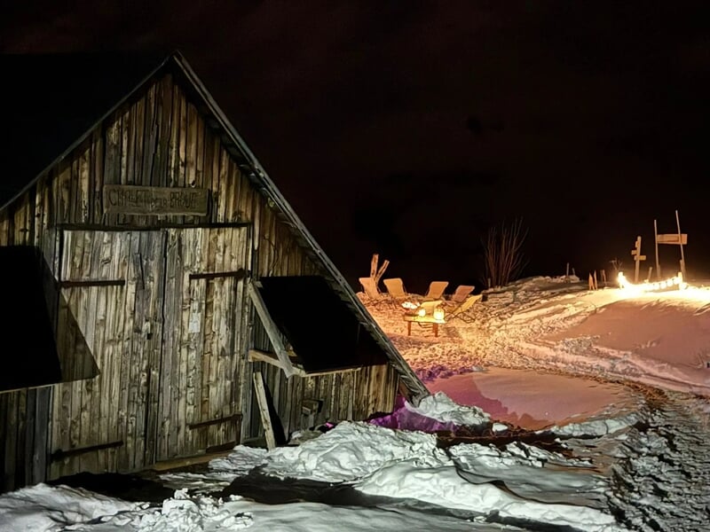Cabane en bois rustique entourée de neige avec un feu de camp et des chaises en bois sous un ciel nocturne