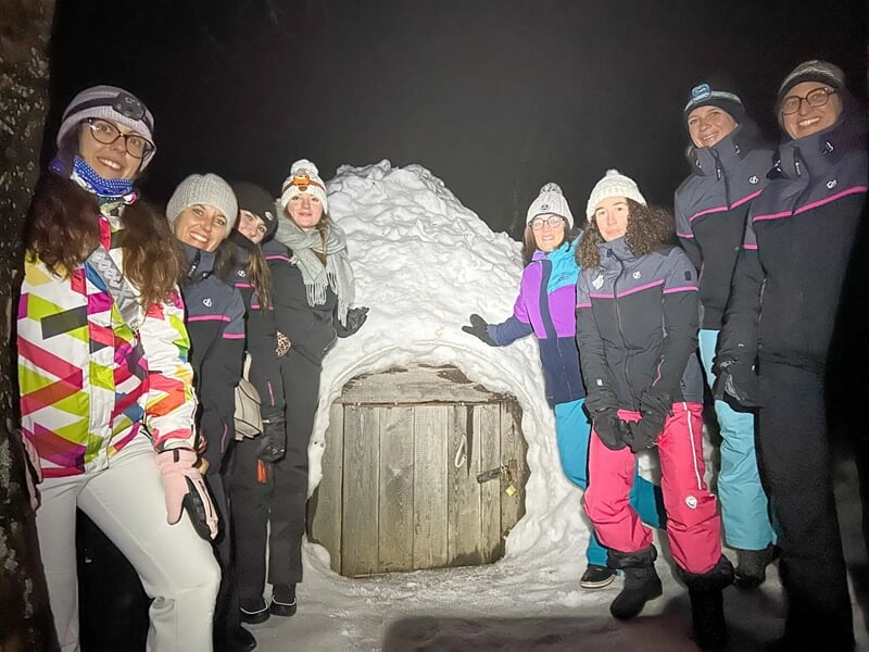 Groupe de sept personnes souriantes posant devant un igloo en neige avec une porte en bois la nuit.