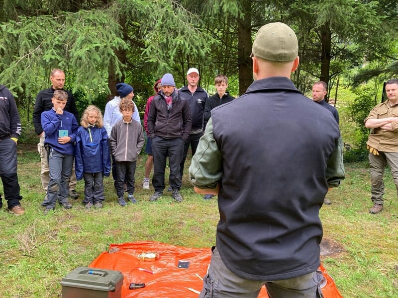 Instructeur de survie de dos portant une casquette et une veste noire devant un groupe attentif en forêt