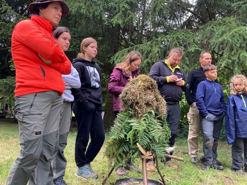 Groupe de personnes de divers âges observant attentivement une structure de survie en forêt, faite de branches et de feuillage