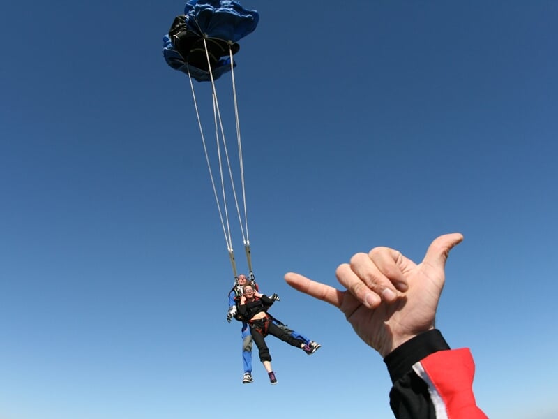 Parachutisme en tandem sous un parachute bleu déployé, avec une main en premier plan faisant un signe