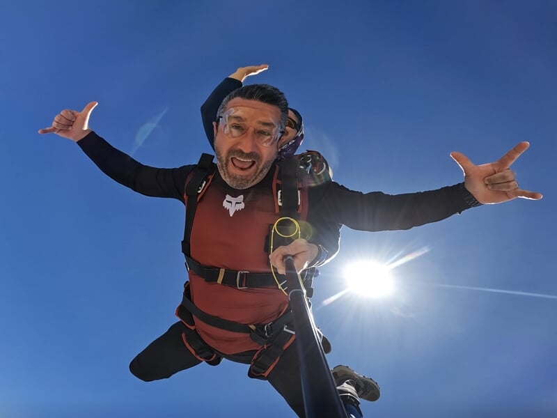 Saut en parachute en tandem avec un homme affichant une expression de joie et d’adrénaline, vêtu d’une combinaison rouge et noire. Il tient une perche à selfie, bras ouverts, avec un ciel bleu éclatan