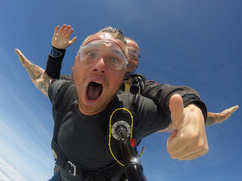 Parachutiste en tandem criant d’excitation, pouce levé, lors d'une chute libre au-dessus des nuages. Équipé d’un harnais et de lunettes de protection, il immortalise son expérience avec une caméra fix