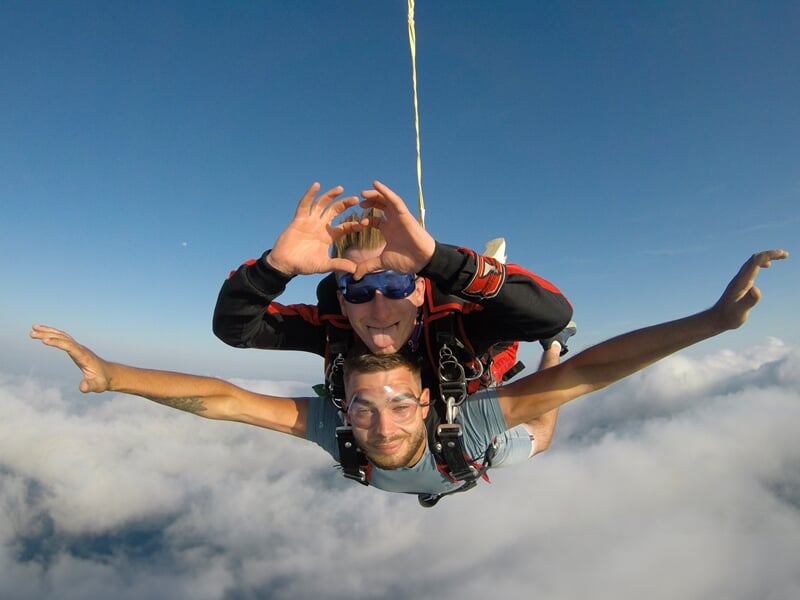 Chute libre en tandem au-dessus des nuages, avec un instructeur souriant formant un cœur avec ses mains. Le parachutiste principal étend les bras, profitant de l'expérience dans un ciel bleu dégagé.