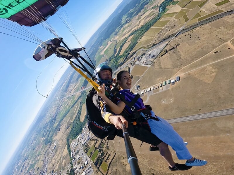 Descente sous parachute biplace avec une voile verte et noire, offrant une vue panoramique sur un paysage de campagne et un aérodrome. Les deux parachutistes sourient, profitant du vol suspendu au-des
