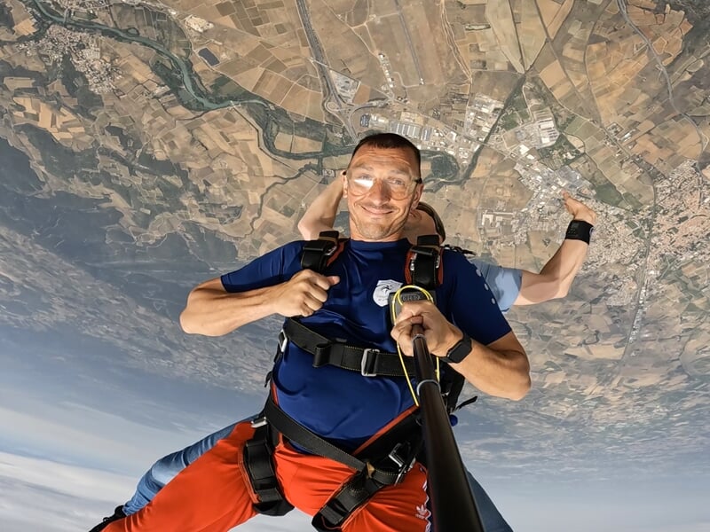 Saut en parachute en tandem avec un homme souriant tenant une perche à selfie, en pleine chute libre au-dessus d’un paysage de plaines et de rivières. L’adrénaline et l’excitation se lisent sur son vi